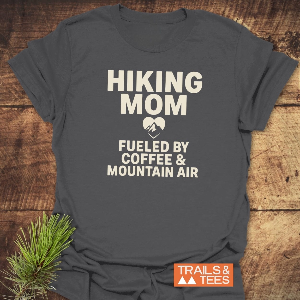 Hiking Mom Mountain Air T-Shirt with Fueled by Coffee & Mountain Air print, laid flat with a pine sprig on a wood surface.