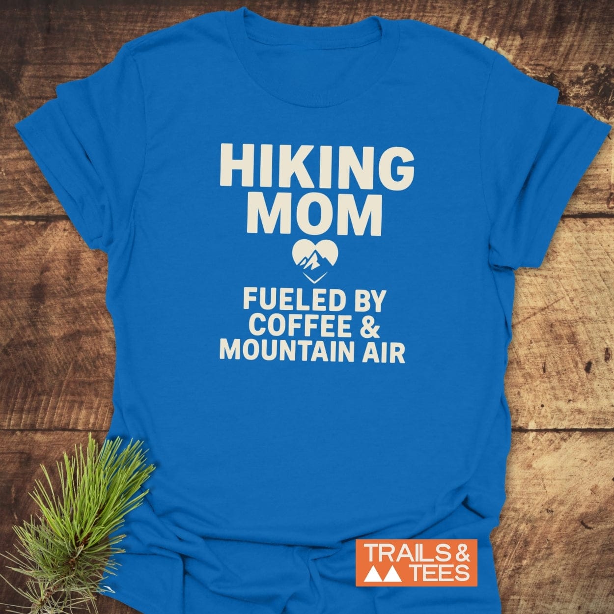 Hiking Mom Mountain Air T-Shirt with Fueled by Coffee & Mountain Air print, featuring durable design, laid flat with pine sprig, emphasizing outdoor adventure theme.