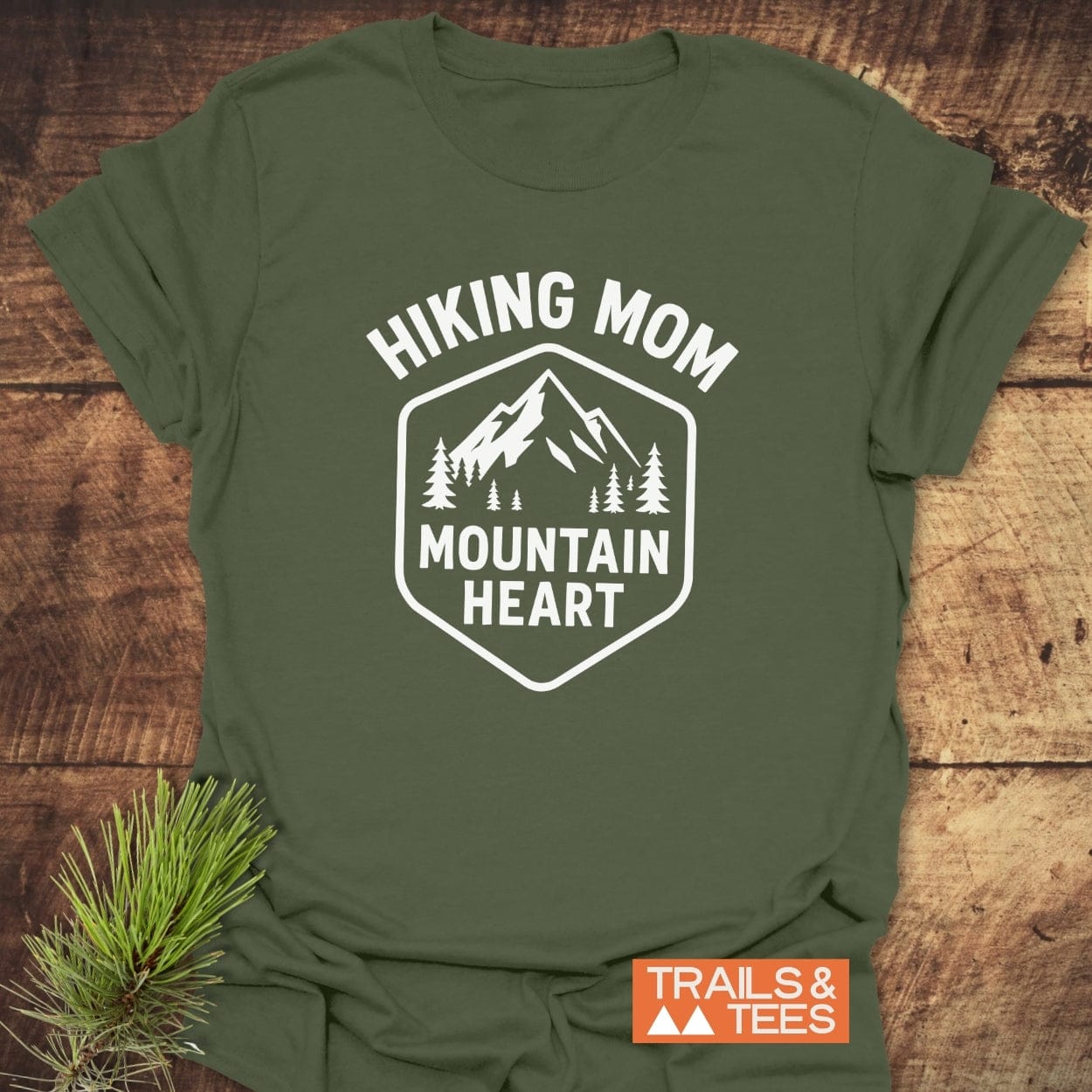 Forest-green Hiking Mom Mountain Heart T-Shirt with mountain badge print, laid on wood. Features soft cotton, durable print, and eco-friendly production.