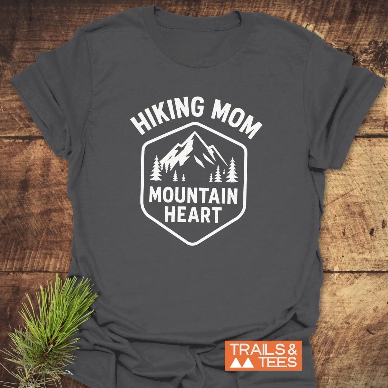 Hiking Mom Mountain Heart T-Shirt featuring a mountain and pine tree graphic, laid on wood with pine branch. Soft, durable cotton, perfect for outdoor enthusiasts.