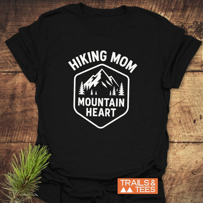 Hiking Mom Mountain Heart T-Shirt featuring a mountain and pine tree graphic, laid on wood. Made from soft cotton, perfect for outdoor enthusiasts.