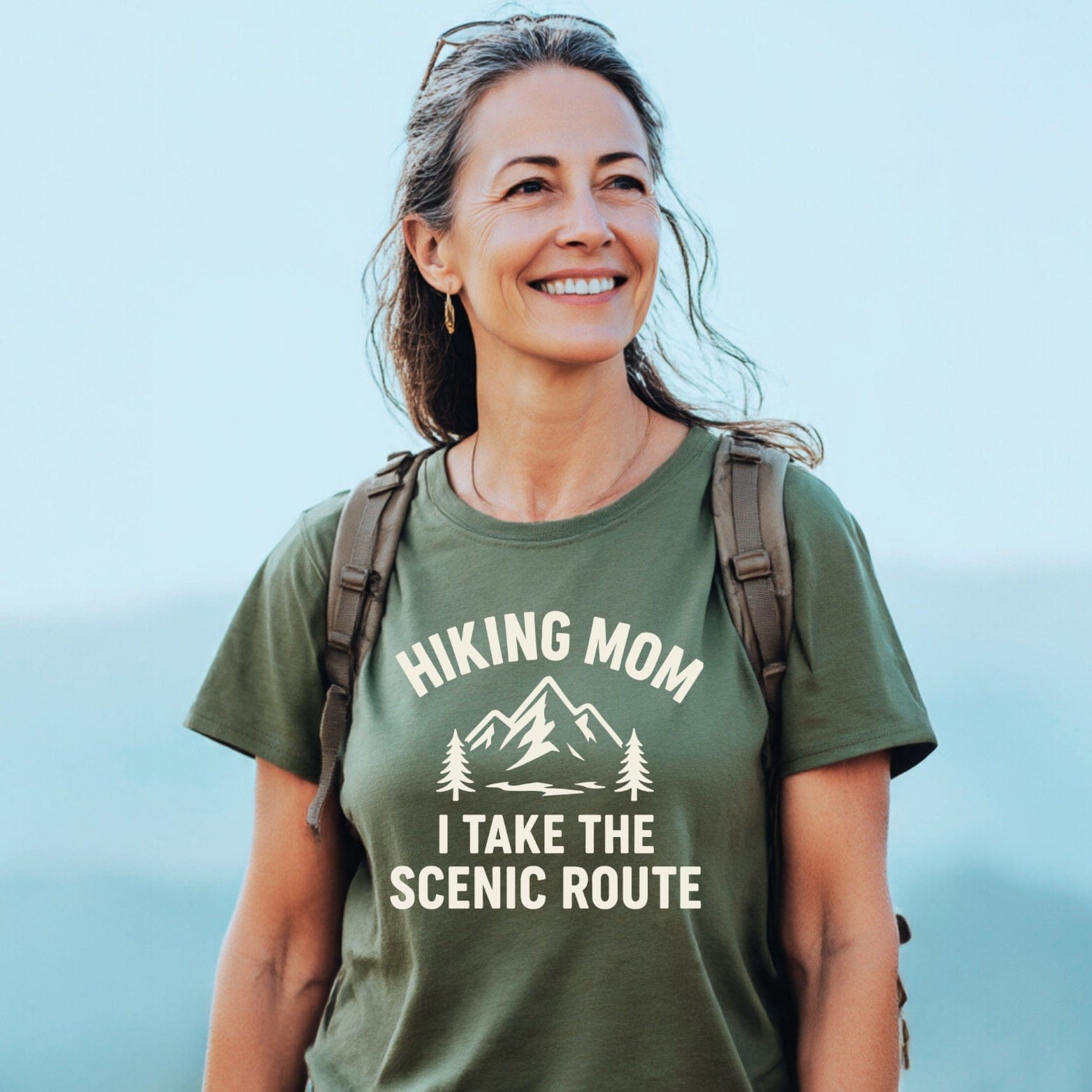 Woman hiking, wearing a Hiking Mom, Scenic Route T-Shirt with mountain graphic, carrying a backpack. T-shirt features durable print, soft cotton, pre-shrunk for fit.