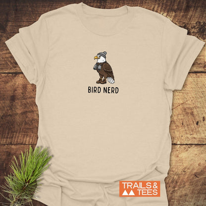 Bird Nerd Funny T-Shirt featuring a cartoon eagle with a beanie and binoculars, emphasizing outdoor adventure and comfort, laid flat on a wooden surface.