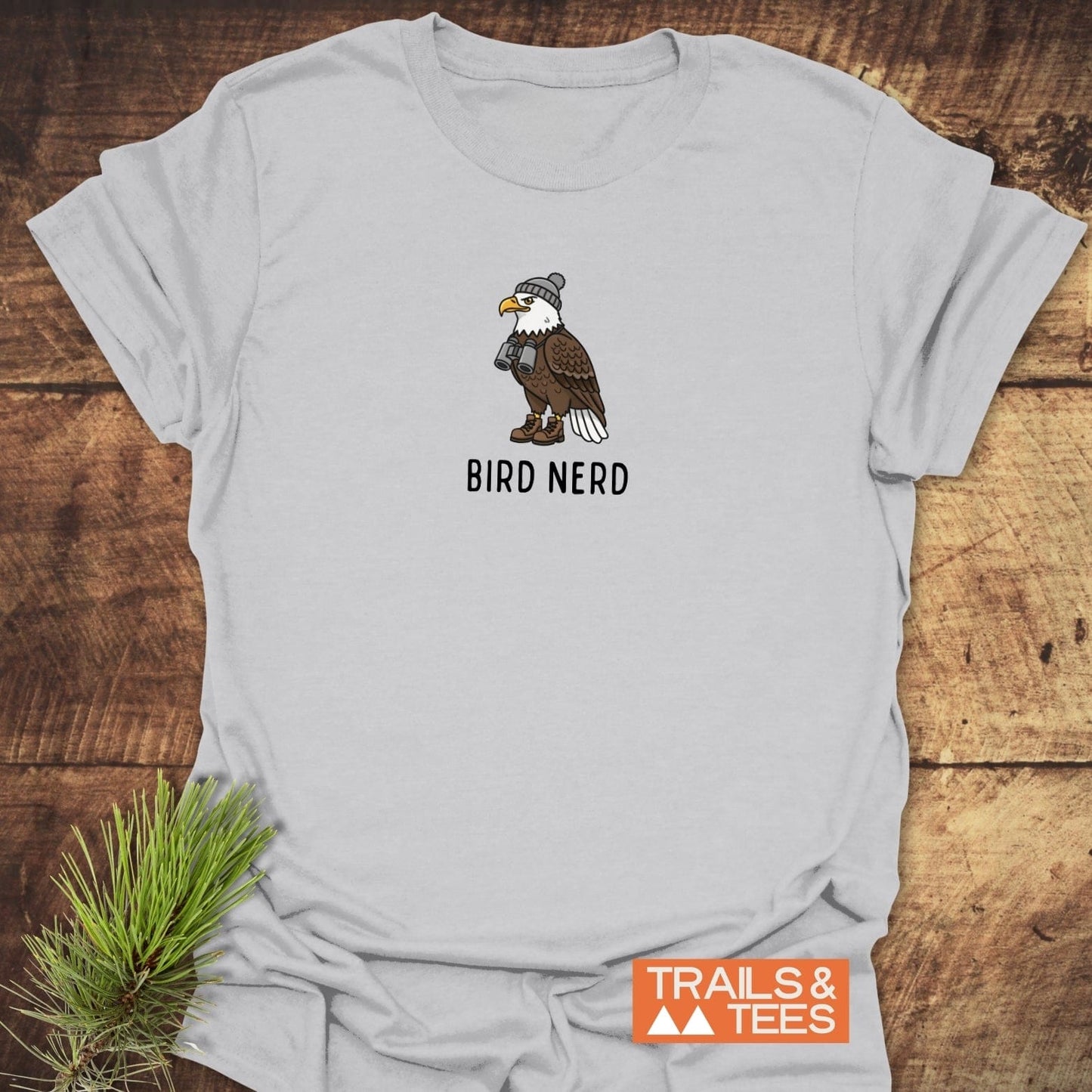 Bird Nerd Funny T-Shirt featuring a cartoon bald eagle with binoculars and boots, laid on wood beside pine, showcasing outdoor adventure spirit.
