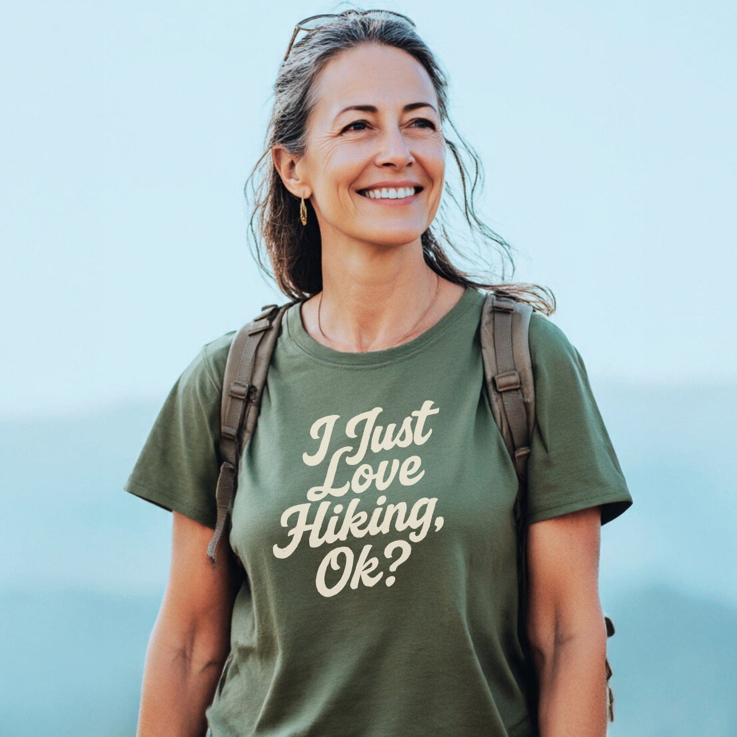 Middle-aged woman outdoors wearing I Just Love Hiking T-shirt, backpack, sunglasses on head; showcasing trail-tested comfort and durable design for outdoor enthusiasts.