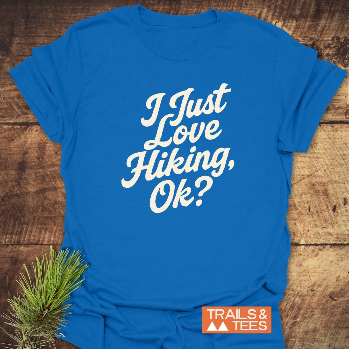 Bright blue I Just Love Hiking T-Shirt with vintage white script, orange TRAILS & TEES logo, laid against wood, featuring outdoorsy pine sprig.
