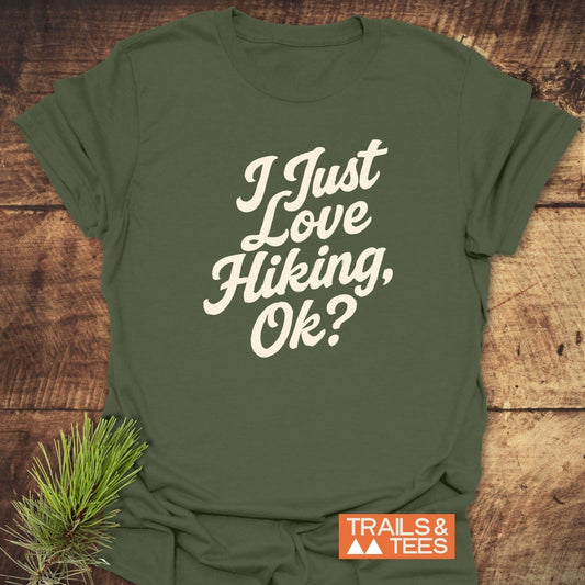 Olive-green I Just Love Hiking T-shirt on wood, showcasing off-white script. Features soft, ring-spun cotton and durable print. Includes TRAILS & TEES logo.