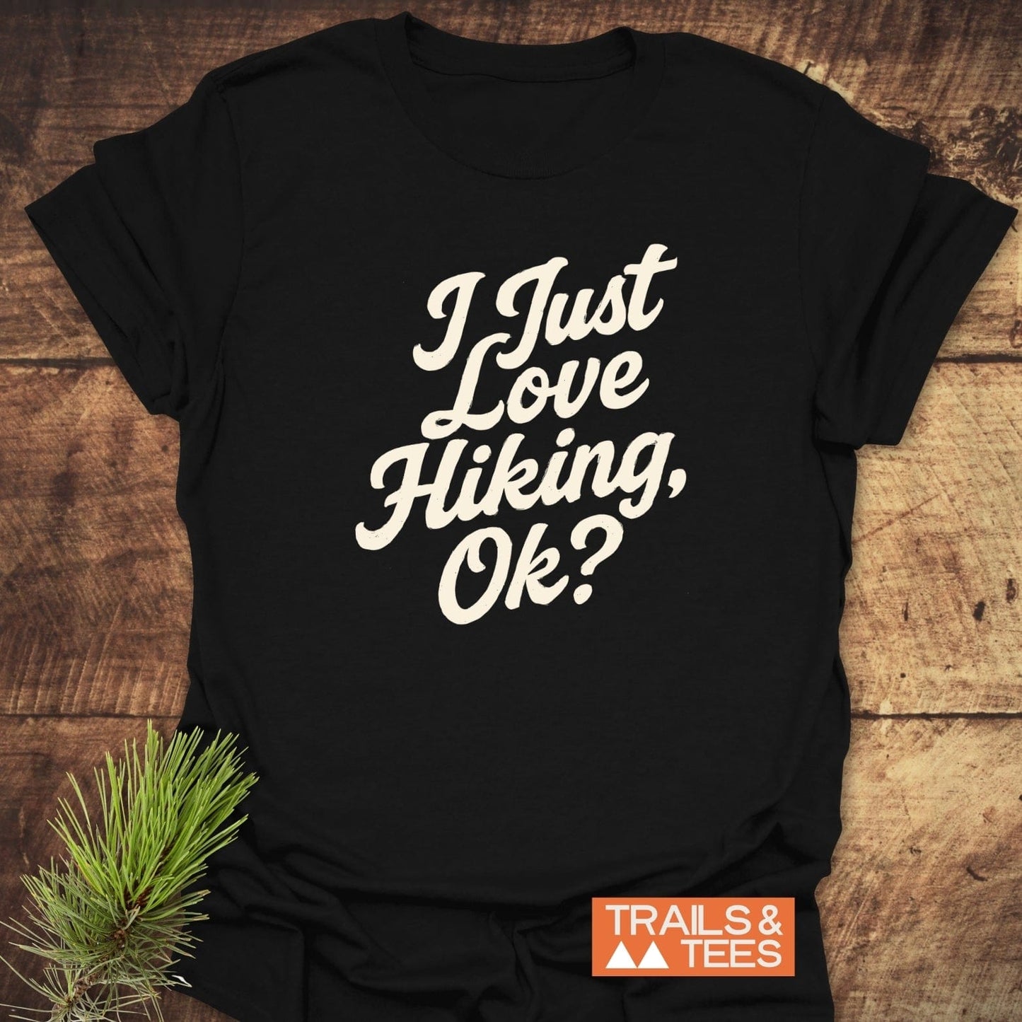 I Just Love Hiking T-Shirt on rustic surface, off-white script, orange 'TRAILS & TEES' logo, pine sprig; soft, ring-spun cotton, pre-shrunk, durable print.