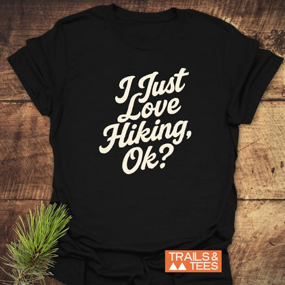 I Just Love Hiking T-Shirt on rustic surface, off-white script, orange 'TRAILS & TEES' logo, pine sprig; soft, ring-spun cotton, pre-shrunk, durable print.