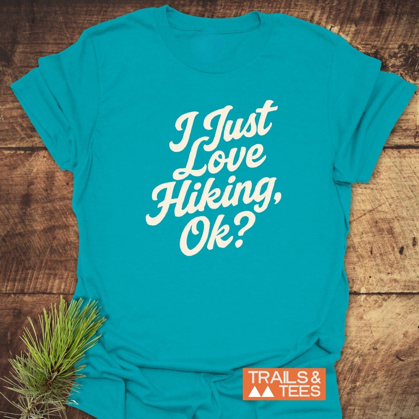 Teal T-shirt with I Just Love Hiking, Ok? printed in white script, featuring the orange TRAILS & TEES logo, complemented by a pine sprig.