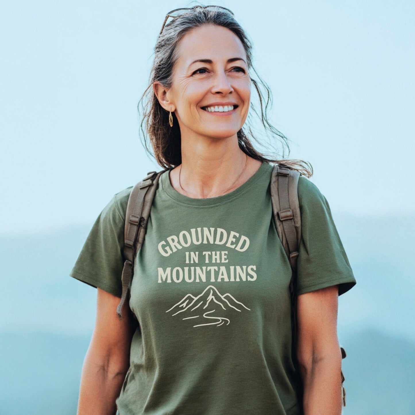 Middle-aged woman outdoors, wearing olive-green Grounded In The Mountains T-Shirt, showcasing its mountain graphic and slogan, with backpack straps visible.