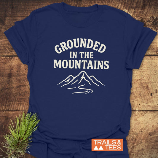 Grounded In The Mountains T-Shirt with mountain art and “TRAILS & TEES” logo, laid on wood, embodies outdoor adventure with durable, comfortable cotton.