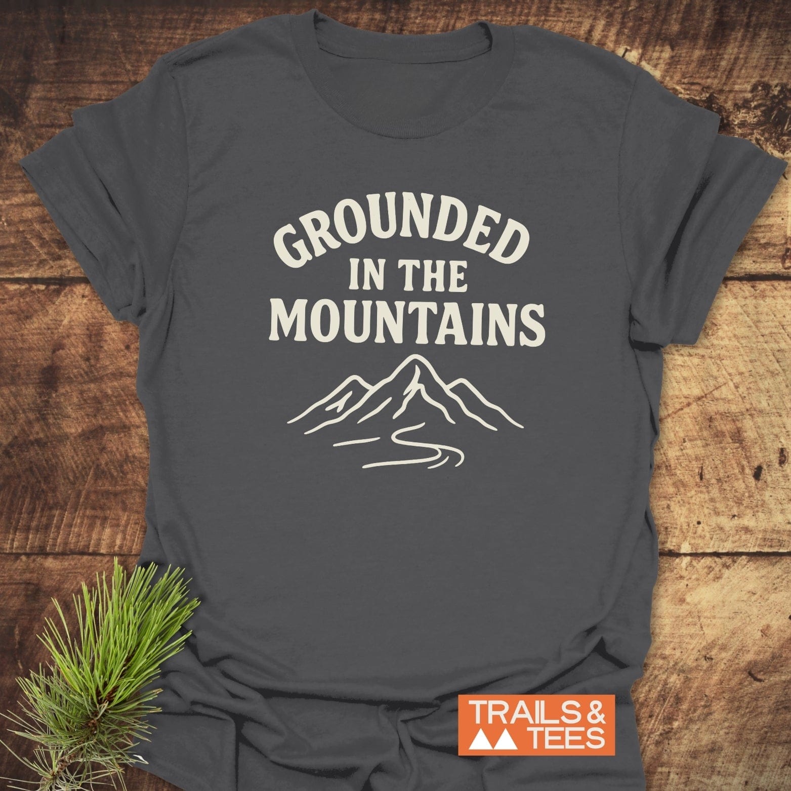 Grounded In The Mountains T-Shirt on wooden surface, featuring mountain graphic and slogan. Soft, ring-spun cotton for comfort, ideal for outdoor enthusiasts.