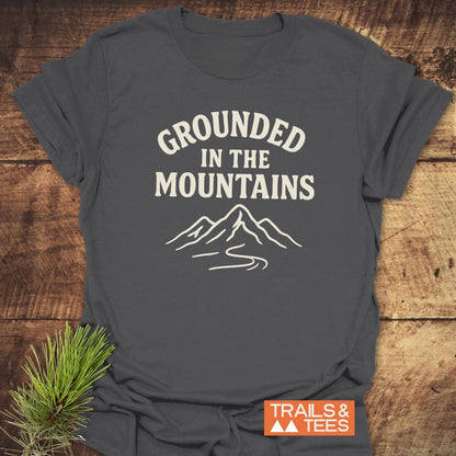 Grounded In The Mountains T-Shirt on wooden surface, featuring mountain graphic and slogan. Soft, ring-spun cotton for comfort, ideal for outdoor enthusiasts.