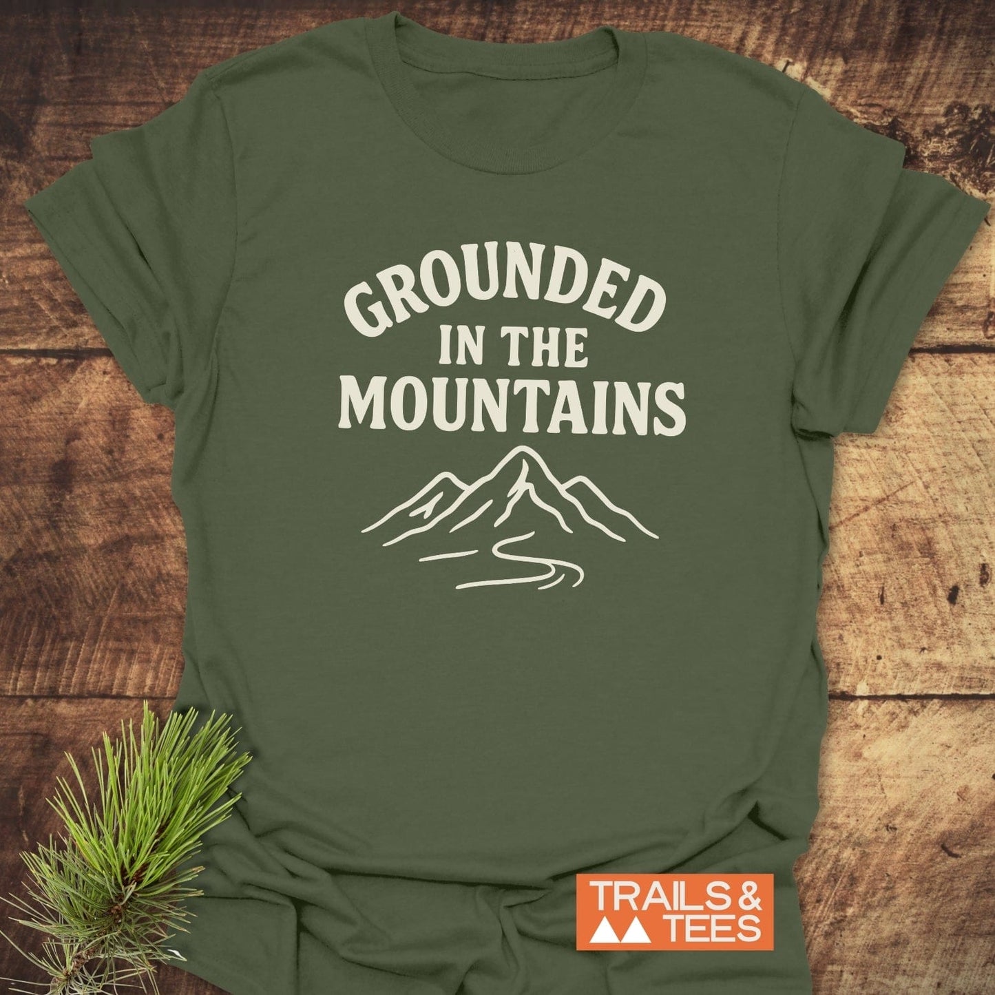 Grounded In The Mountains T-Shirt on wood, featuring off-white mountain design. Soft, ring-spun cotton, pre-shrunk, durable print. Trails & Tees logo visible.