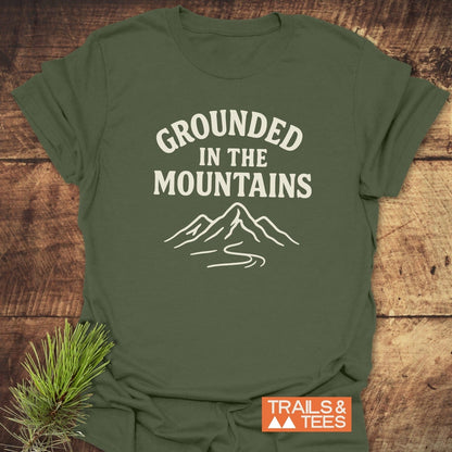 Grounded In The Mountains T-Shirt on wood, featuring off-white mountain design. Soft, ring-spun cotton, pre-shrunk, durable print. Trails & Tees logo visible.