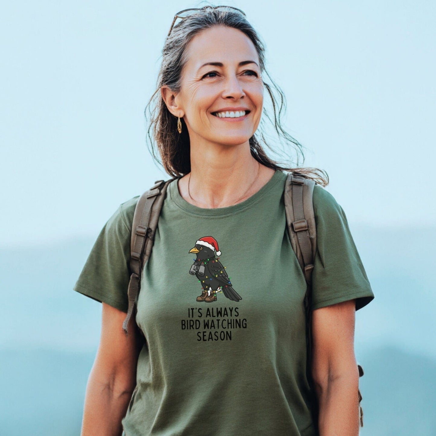 A woman outdoors wears the Always Bird Watching... T-Shirt featuring a festive bird design. The tee offers comfort and durability, ideal for adventure lovers.