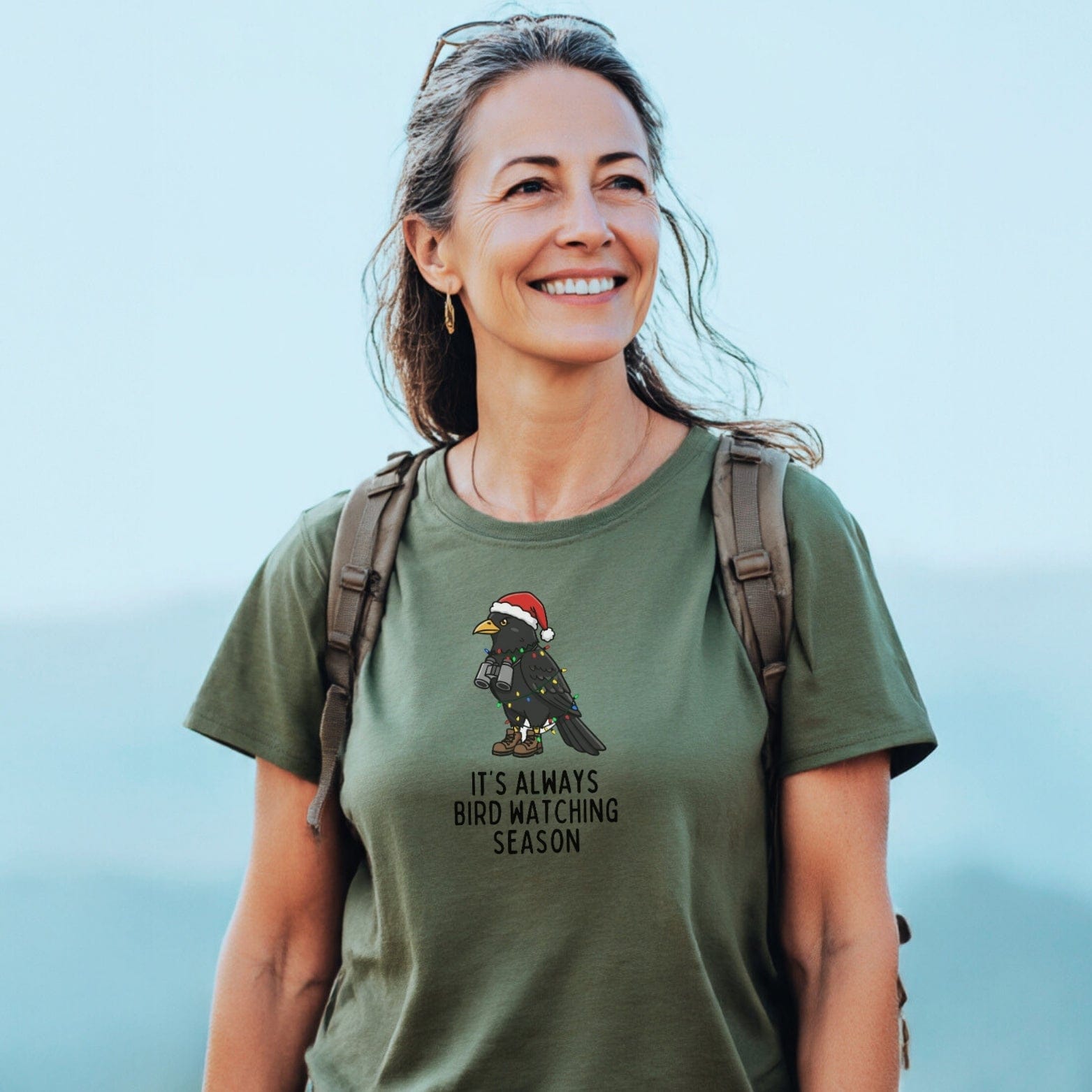 A woman outdoors wears the Always Bird Watching... T-Shirt featuring a festive bird design. The tee offers comfort and durability, ideal for adventure lovers.