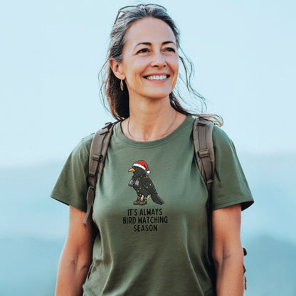 A woman outdoors wears the Always Bird Watching... T-Shirt featuring a festive bird design. The tee offers comfort and durability, ideal for adventure lovers.