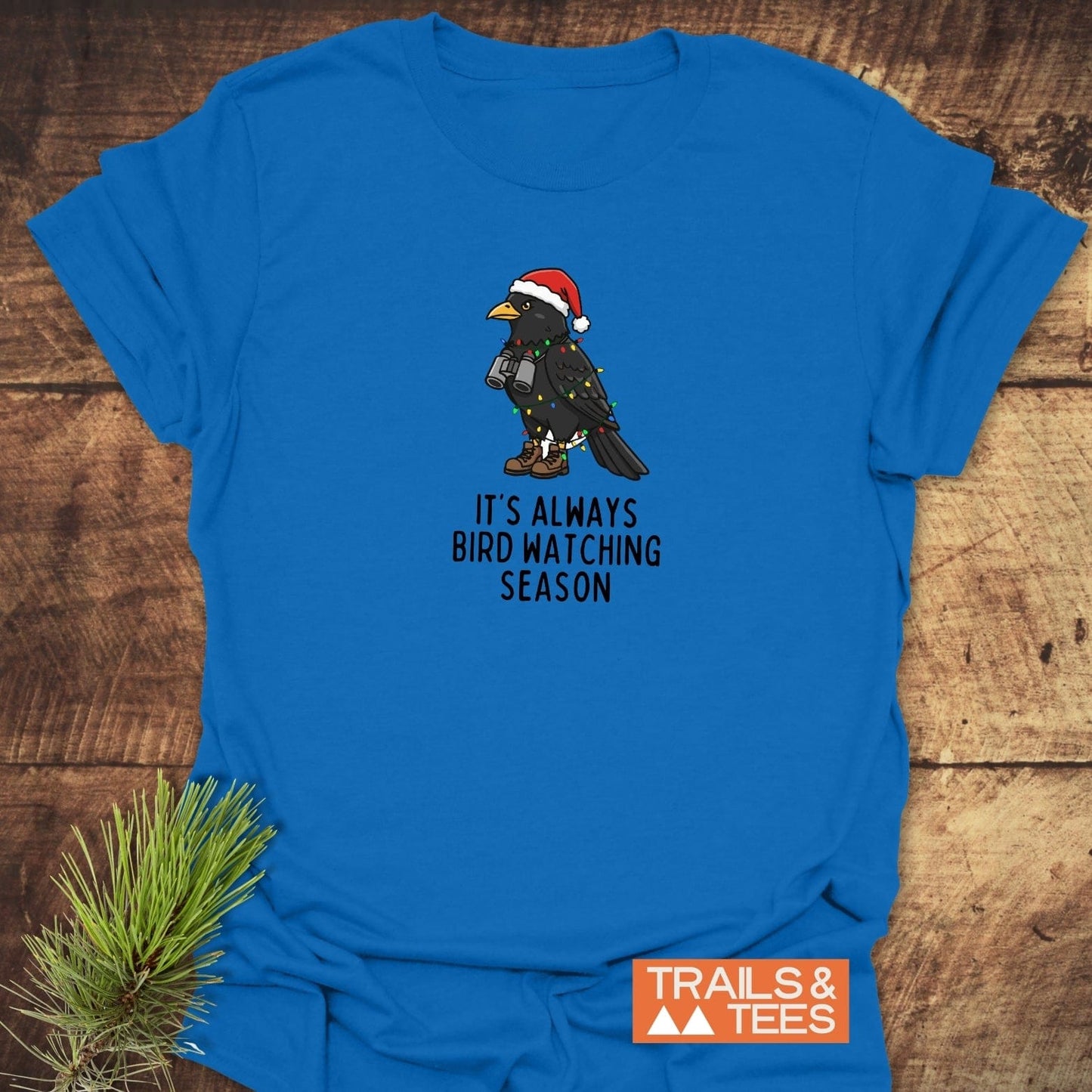 Always Bird Watching... T-Shirt with holiday-themed blackbird design, featuring Santa hat, lights, hiking boots, and binoculars; slogan promotes year-round bird watching.
