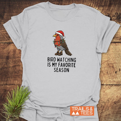 Bird Watching Season T-Shirt featuring a cartoon robin in hiking boots with Christmas lights and a Santa hat, on a wooden surface with pine sprig beside it.