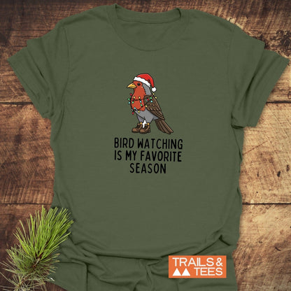 Bird Watching Season T-Shirt with cartoon robin in Santa hat, hiking boots, and Christmas lights on olive-green fabric, laid on a wooden surface.