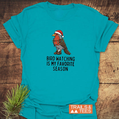 Bird Watching Season T-Shirt featuring a cartoon bird in a Santa hat and boots, wrapped in lights, on soft, pre-shrunk cotton. Trails & Tees logo visible.