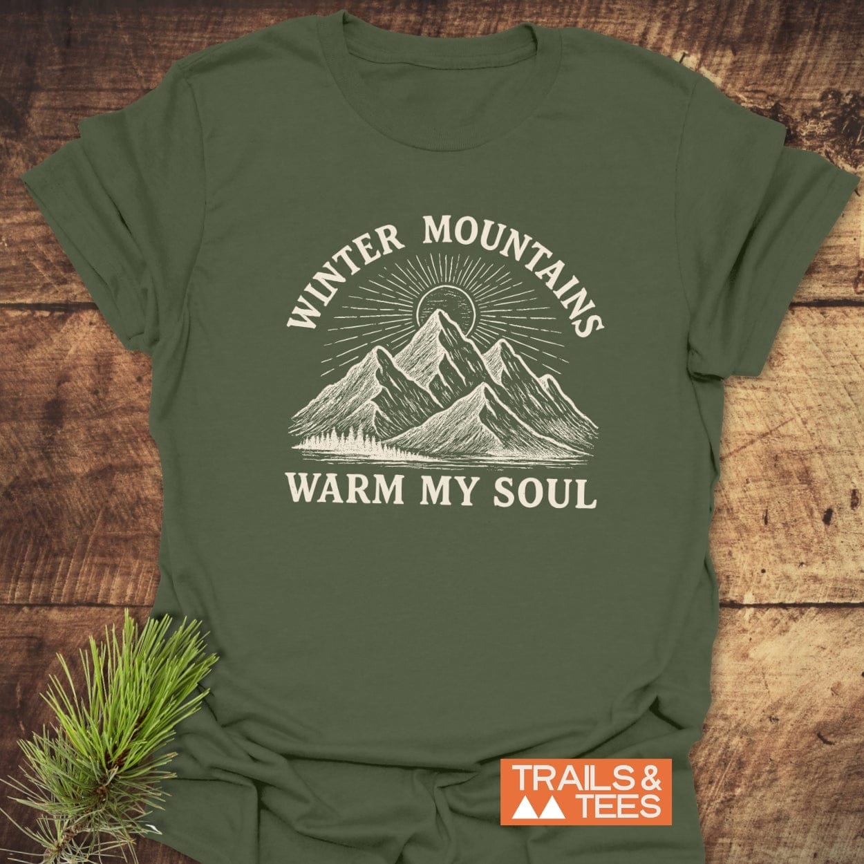 Forest-green Winter Mountains T-Shirt with a hand-drawn mountain and sunburst design, slogan “WINTER MOUNTAINS WARM MY SOUL,” laid on wood, with a pine twig and “TRAILS & TEES” label visible.