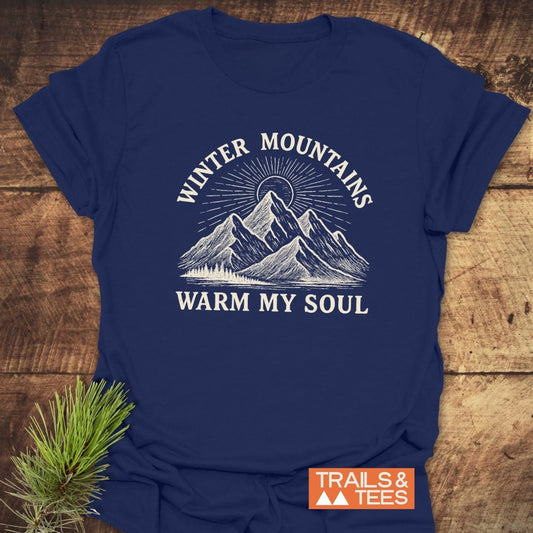 Navy-blue Winter Mountains T-Shirt featuring a white, hand-drawn illustration of snow-capped mountains with a sunburst. Includes WINTER MOUNTAINS and WARM MY SOUL text, plus a TRAILS & TEES logo.