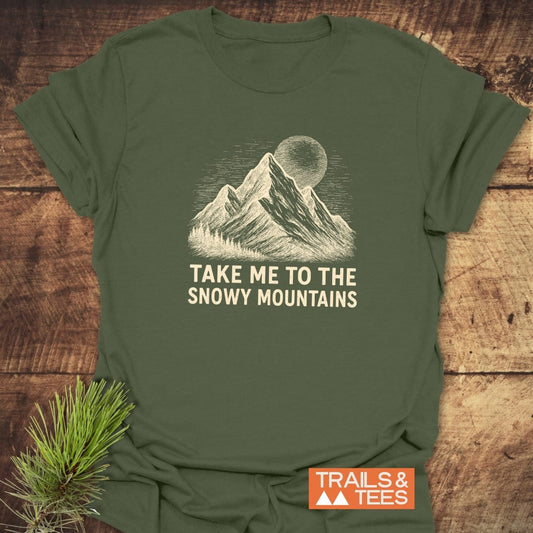Snowy Mountains T-Shirt featuring a mountain range and sun/moon design with TAKE ME TO THE SNOWY MOUNTAINS slogan, laid flat with a pine sprig, Trails & Tees logo visible.