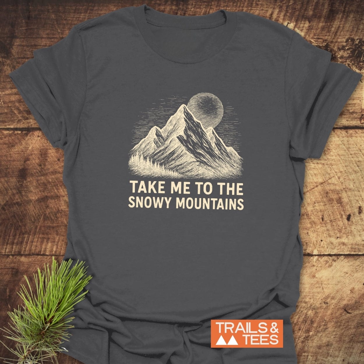 Snowy Mountains T-Shirt featuring a hand-etched mountain summit illustration with rising sun/moon, slogan “TAKE ME TO THE SNOWY MOUNTAINS,” laid flat on a wooden surface, branded “TRAILS & TEES.”