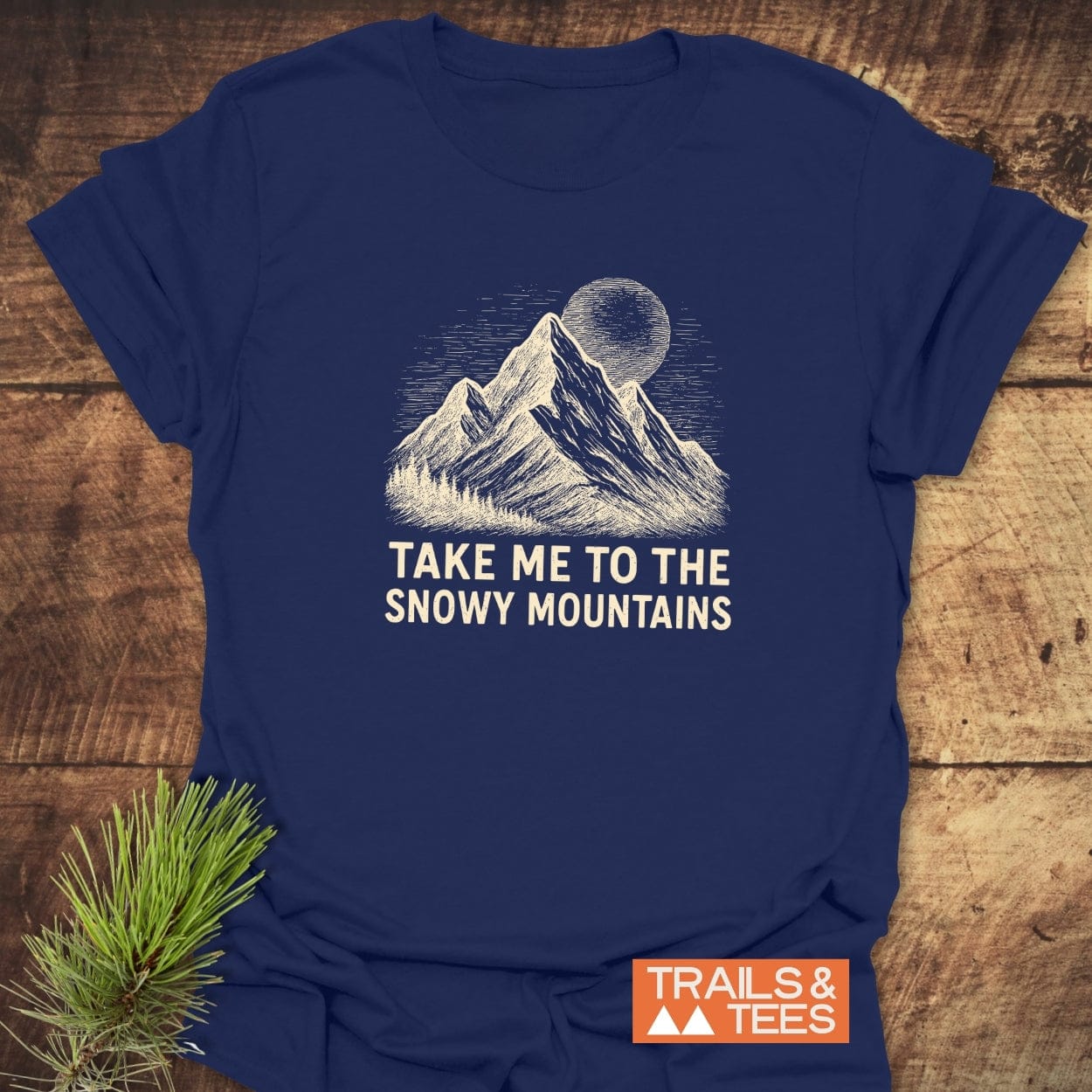 Snowy Mountains T-Shirt featuring a hand-drawn mountain and moon illustration with TAKE ME TO THE SNOWY MOUNTAINS text, laid on wood, highlighting outdoor adventure spirit and comfort.