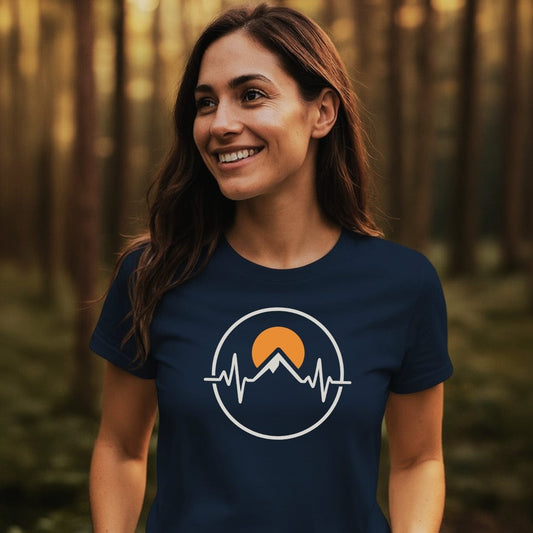Young woman in a forest wears the Heartbeat Mountain T-Shirt, featuring a mountain peak heartbeat graphic and rising sun. The soft, durable tee embodies adventure and outdoor passion.