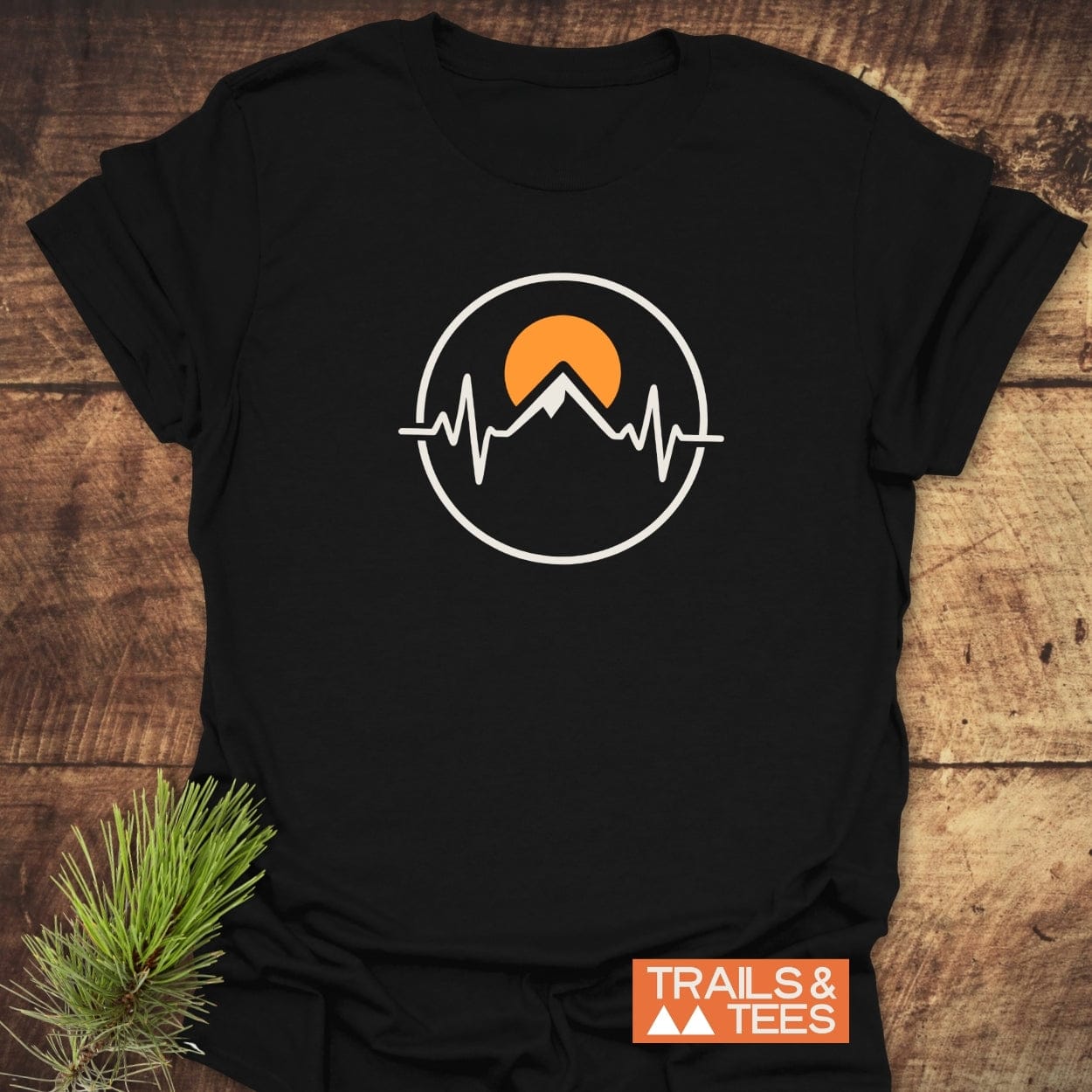 Heartbeat Mountain T-Shirt: Black tee with an orange semicircle sun and mountain ECG line logo. Orange TRAILS & TEES label and pine sprig accent on a wooden background.