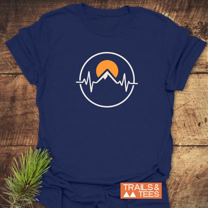 Heartbeat Mountain T-Shirt laid on wood, featuring an emblem of a mountain peak with a heartbeat line and sun. Includes TRAILS & TEES label, ideal for outdoor enthusiasts.