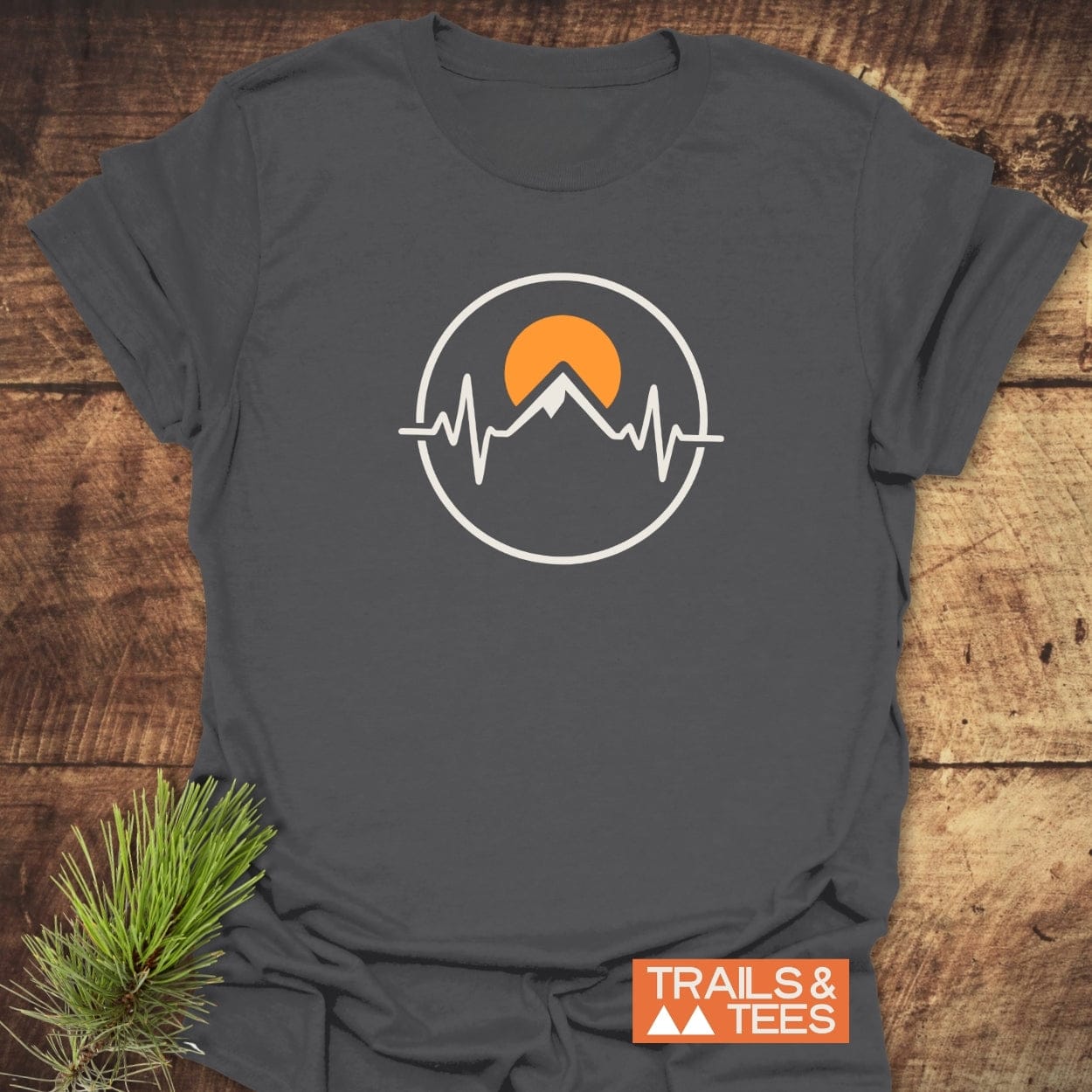 Heartbeat Mountain T-Shirt featuring a circular graphic with a stylized mountain peak, orange sun, and EKG line motif. Includes TRAILS & TEES brand tag with mountain icons. Laid on wooden surface.