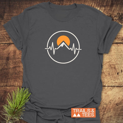 Heartbeat Mountain T-Shirt featuring a circular graphic with a stylized mountain peak, orange sun, and EKG line motif. Includes TRAILS & TEES brand tag with mountain icons. Laid on wooden surface.