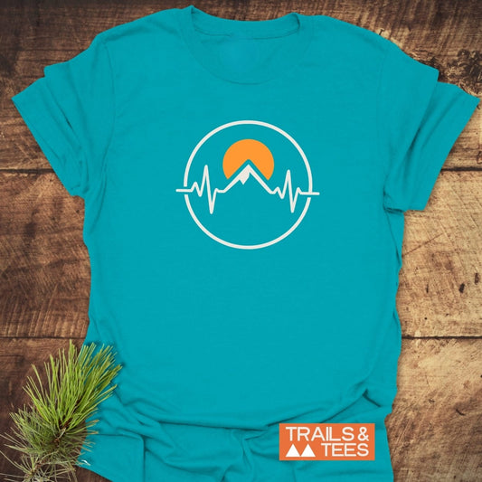Heartbeat Mountain T-Shirt featuring a white circle with a mountain and EKG line graphic, orange sun, on a turquoise tee. Styled with a pine sprig, showcasing adventure-inspired design.