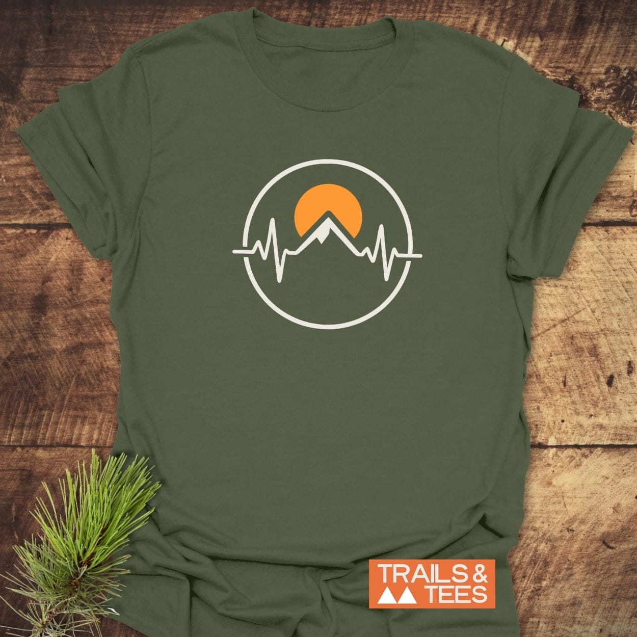 Heartbeat Mountain T-Shirt featuring a mountain and EKG line emblem, with an orange sun, on a forest-green T-shirt. Laid on wood, emphasizing outdoor adventure and comfort.