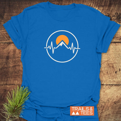 Heartbeat Mountain T-Shirt with a white circular logo of a mountain peak and EKG line, featuring an orange sun. Displayed on a wooden surface with a TRAILS & TEES label and pine sprig.