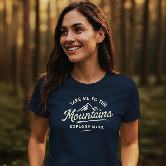 A woman smiles in a forest wearing the Take Me To The Mountains T-Shirt, featuring a mountain graphic and motivational text, ideal for outdoor enthusiasts seeking comfort and style.