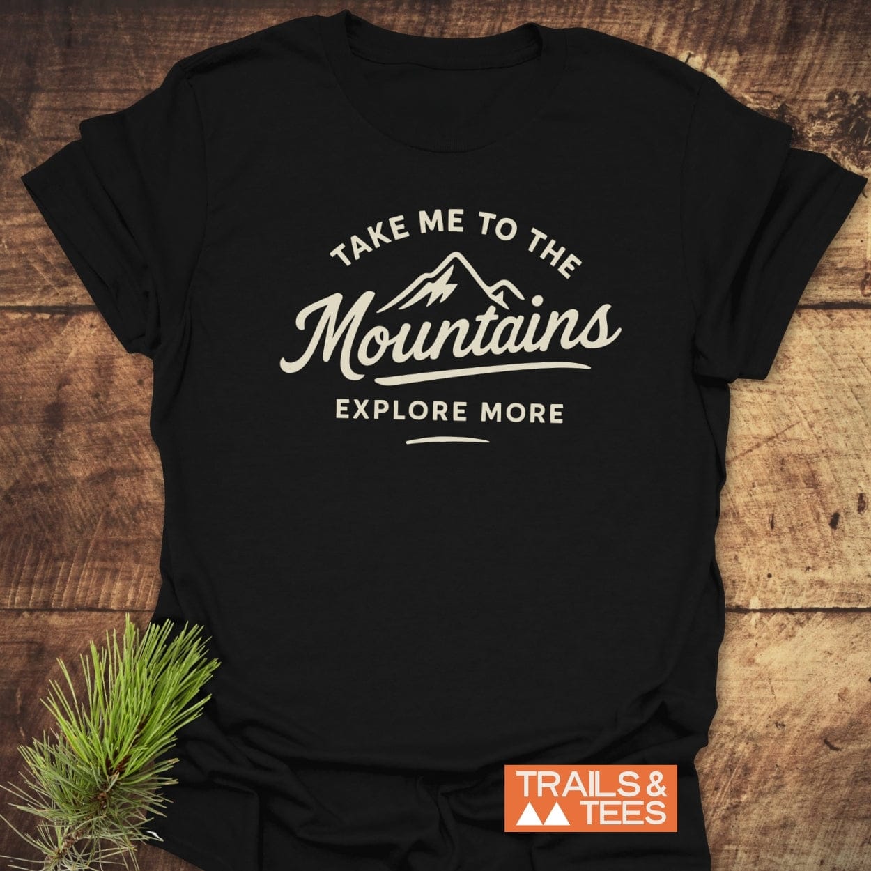 Take Me To The Mountains T-Shirt featuring a mountain graphic and slogans, laid on wood. Includes “TRAILS & TEES” logo and pine branch accent, emphasizing outdoor adventure and comfort.