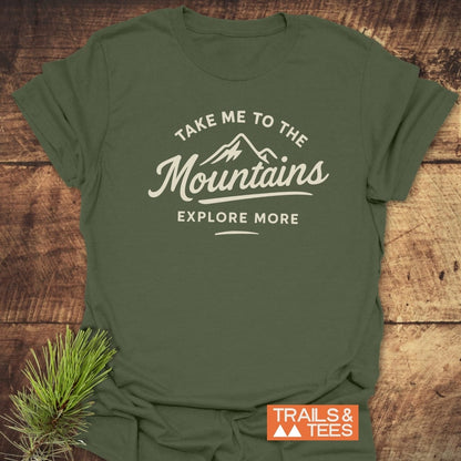 Take Me To The Mountains T-Shirt featuring a mountain graphic and slogan on a forest-green fabric, laid on wood, with durable prints and a small evergreen sprig accent.
