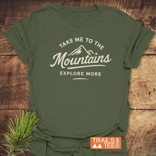 Take Me To The Mountains T-Shirt featuring a mountain graphic and slogan on a forest-green fabric, laid on wood, with durable prints and a small evergreen sprig accent.