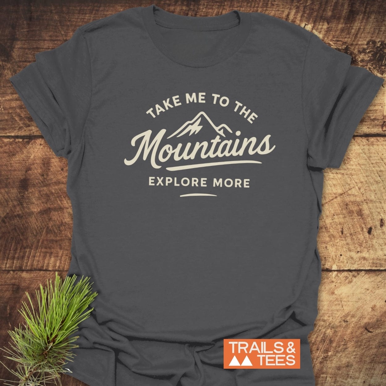 Take Me To The Mountains T-Shirt features a cream mountain graphic with adventurous text, laid flat on wood, alongside a pine sprig and orange TRAILS & TEES label, highlighting outdoor spirit.