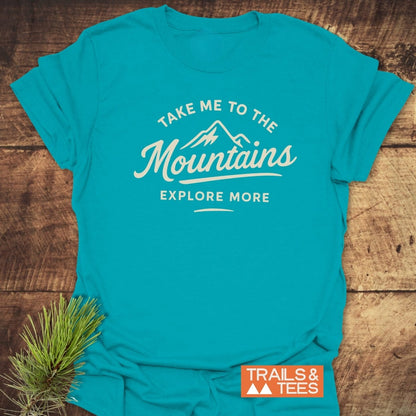 Take Me To The Mountains T-Shirt featuring a white mountain icon and slogan on a teal tee, laid on wooden surface with pine sprig, showcasing Trails & Tees logo, highlighting outdoor adventure spirit.