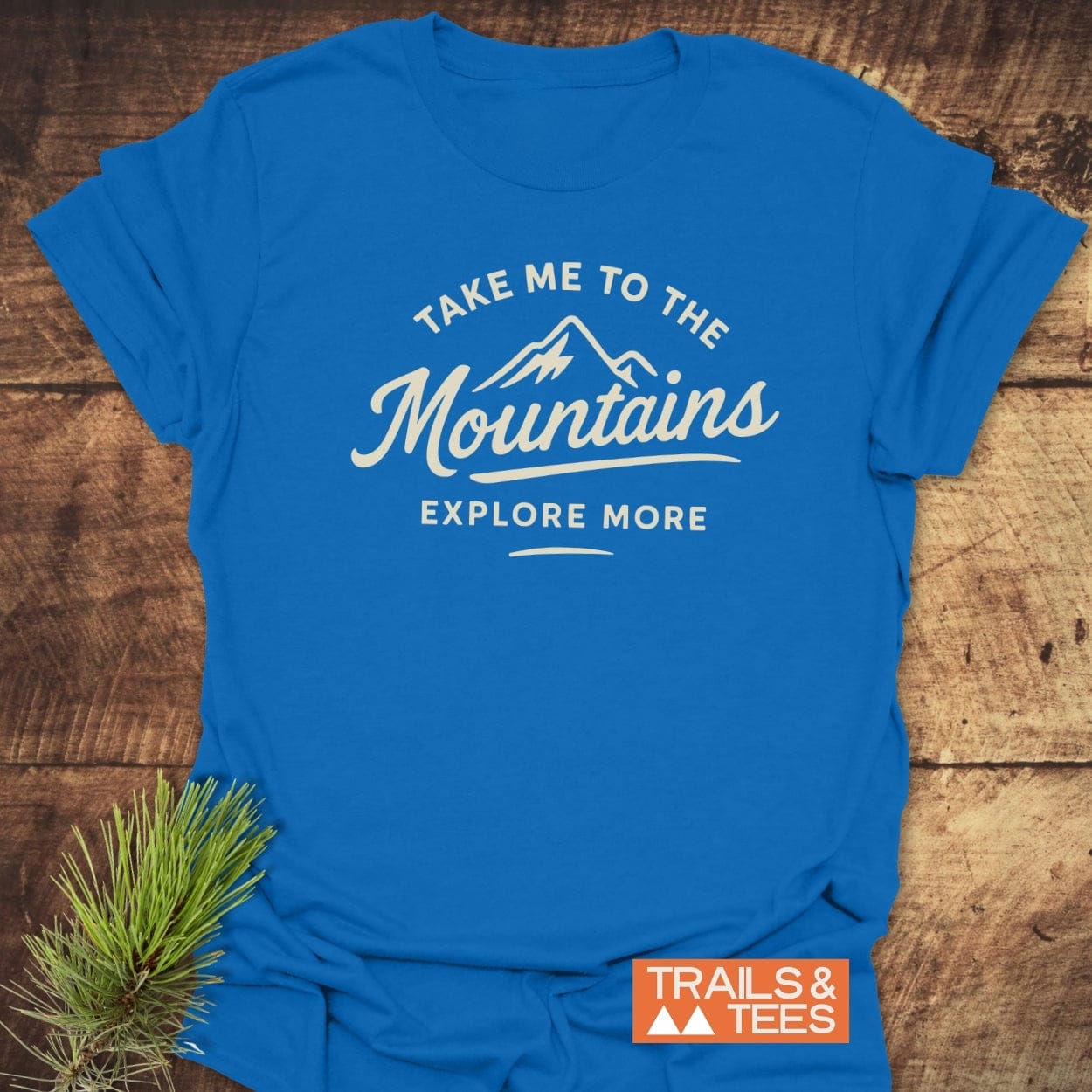 Take Me To The Mountains T-Shirt featuring a stylized mountain graphic and slogans printed on bright blue fabric, laid on wood with a pine sprig, embodying adventure and outdoor spirit.