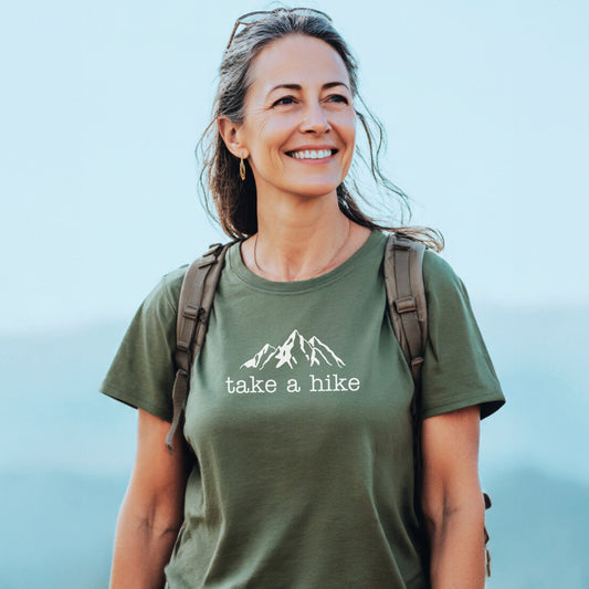 Middle-aged woman outdoors wearing the Take A Hike T-Shirt with a white mountain graphic and “take a hike” text, complemented by backpack straps, ready for an adventure.