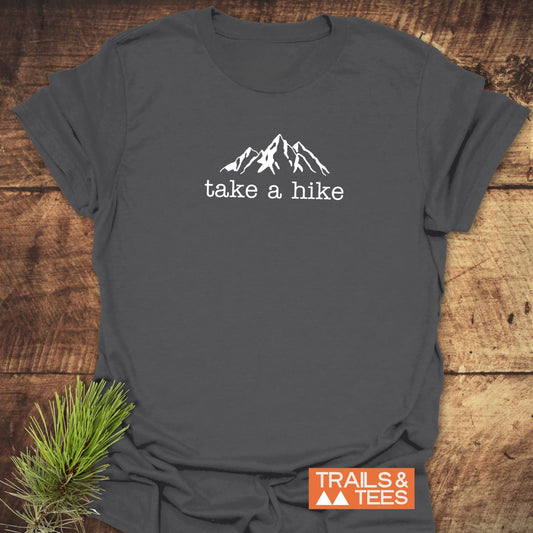 Take A Hike T-Shirt featuring a white mountain peaks line-drawing and take a hike text on dark gray cotton, laid on wood. Includes TRAILS & TEES logo and pine sprig accent.