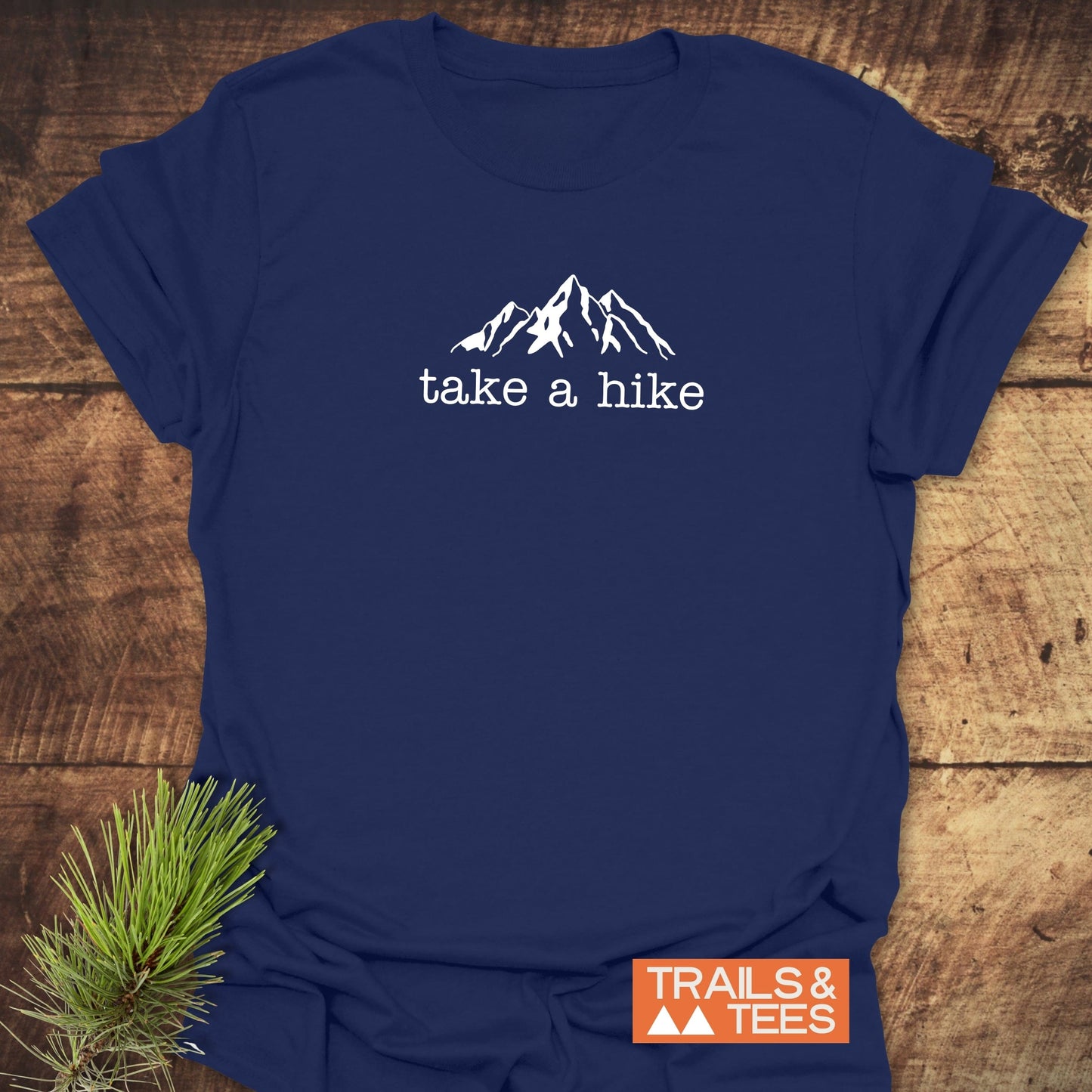 Take A Hike T-Shirt featuring a white mountain graphic and take a hike text on navy-blue fabric, styled with a pine twig, showcasing its outdoor-inspired design and comfort.